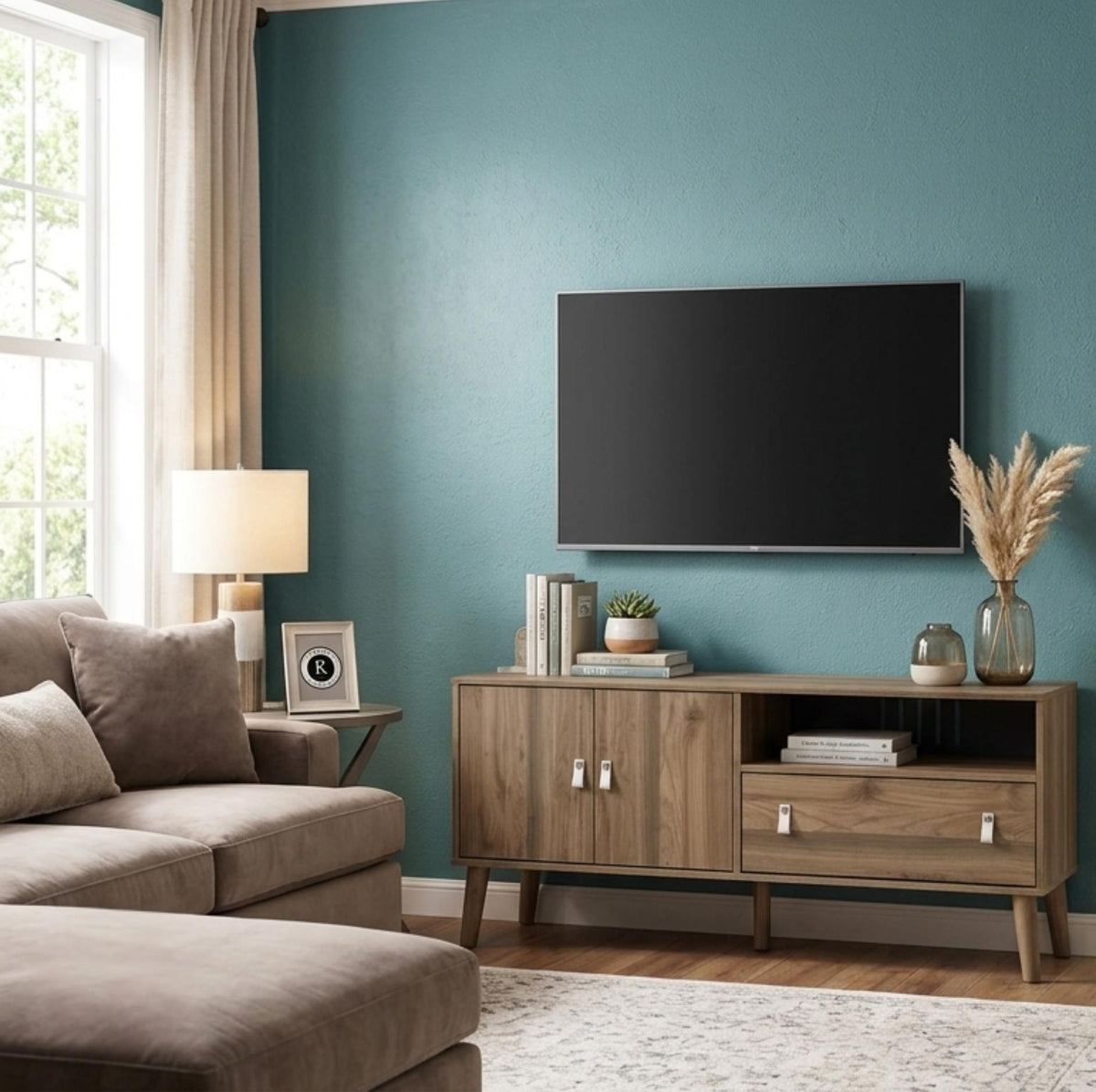 A modern living room scene featuring a light brown wood TV stand console beneath a wall-mounted flat-screen television. The room has a textured teal-blue wall, a beige sofa, and a window with beige curtains.