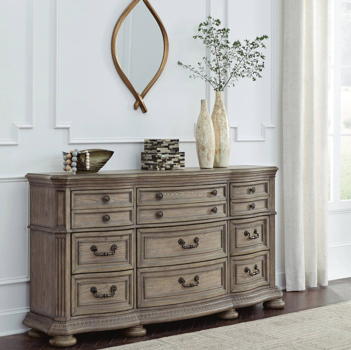 A rustic gray-brown wooden dresser with multiple drawers and dark metal handles, styled in a bright room with decorative vases, a bowl, and a mirror on top.