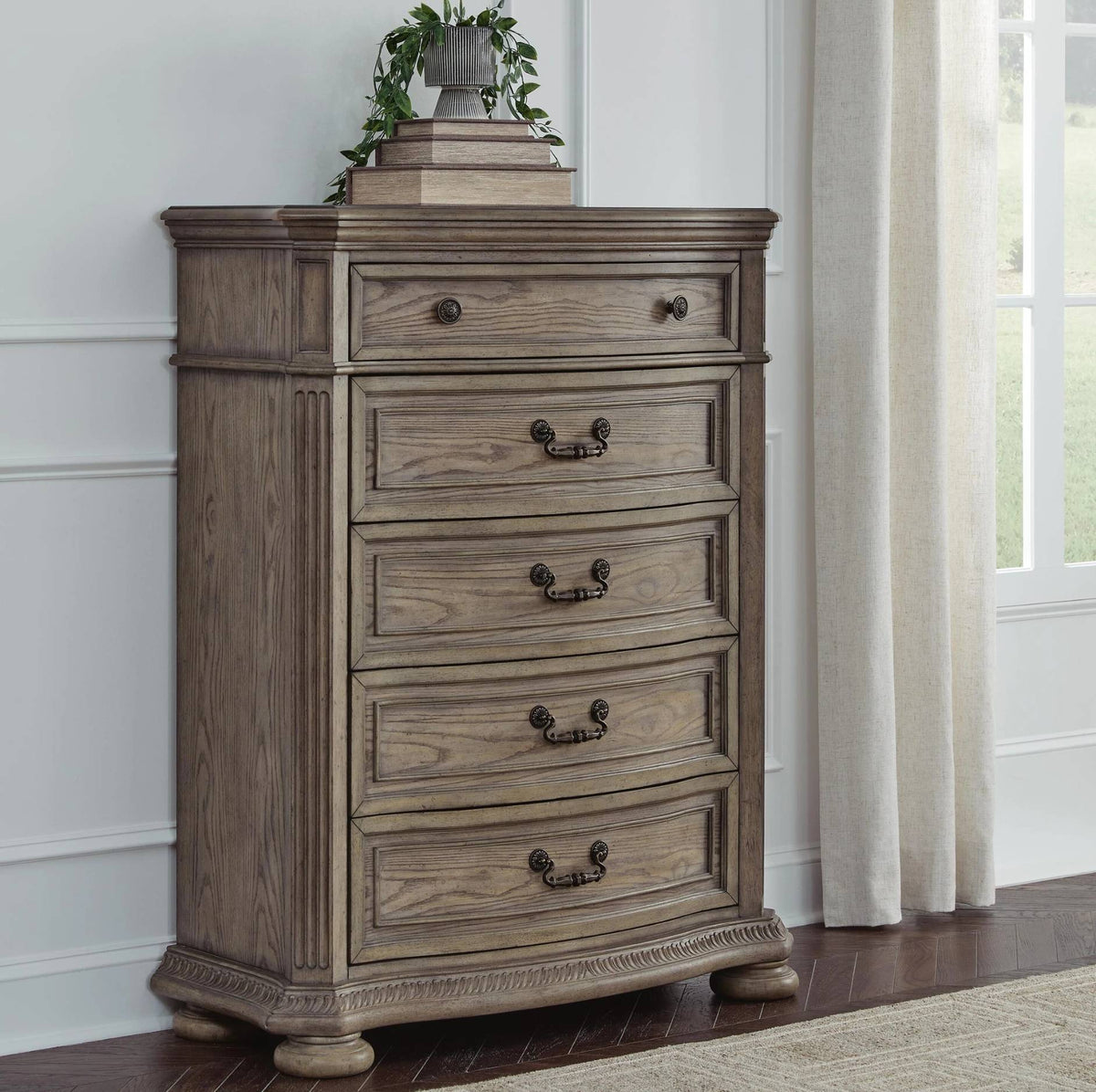 A rustic five-drawer wooden chest of drawers in a weathered gray-brown finish, styled in a room setting next to a window with curtains and topped with a small stack of books and a potted plant.