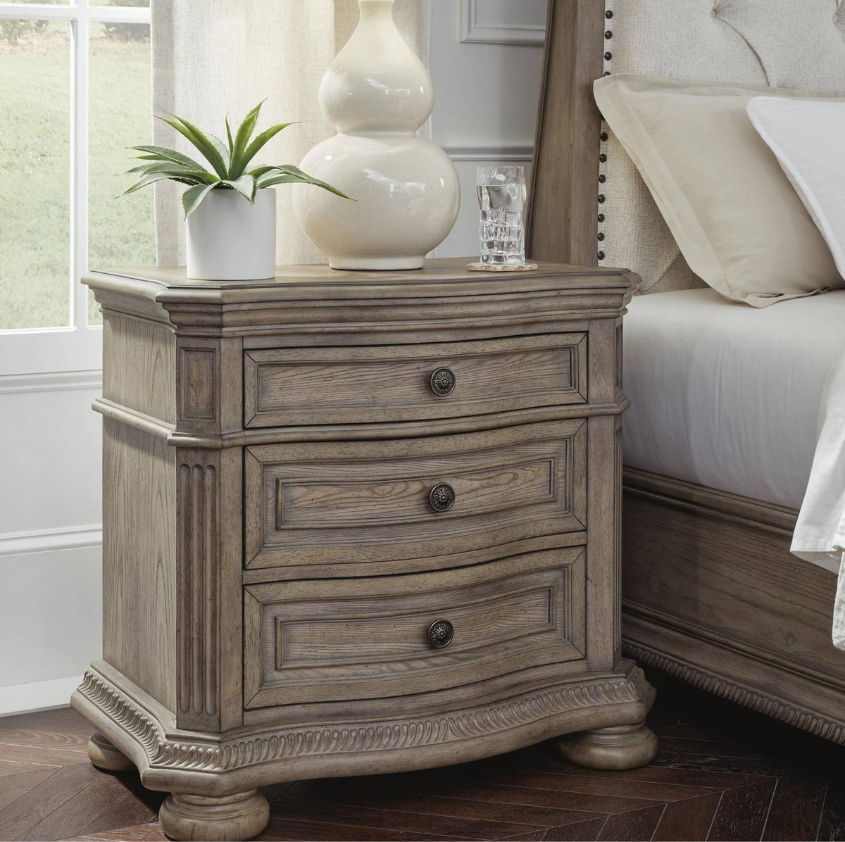 A rustic gray-brown wooden nightstand with three drawers and dark metal knobs, styled next to a bed with neutral bedding and decorative items on top.
