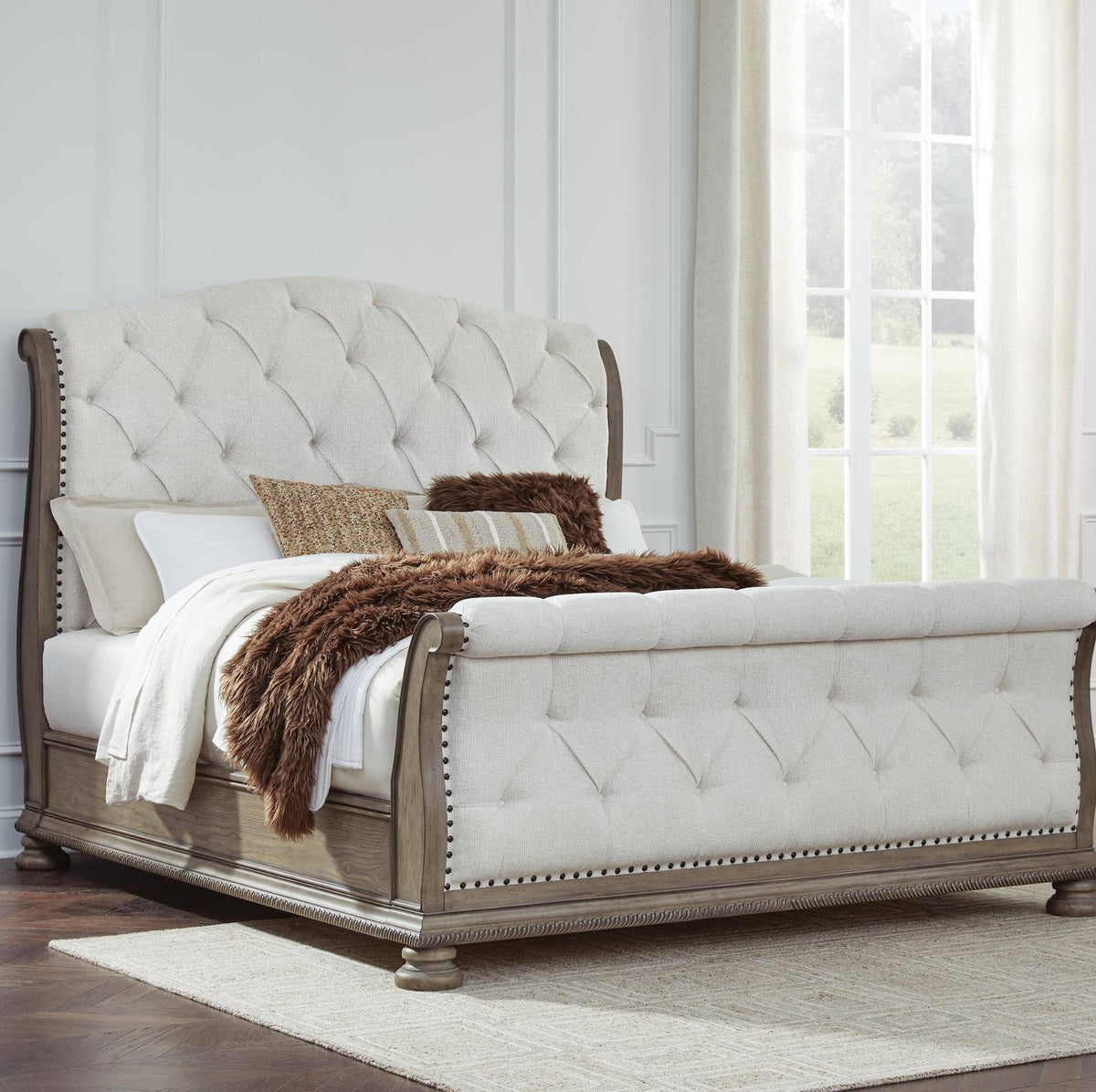 A classic sleigh bed with a tufted cream upholstered headboard and footboard, supported by a weathered gray-brown wood frame with nailhead trim, styled with white linens and brown throw pillows in a bright bedroom setting.