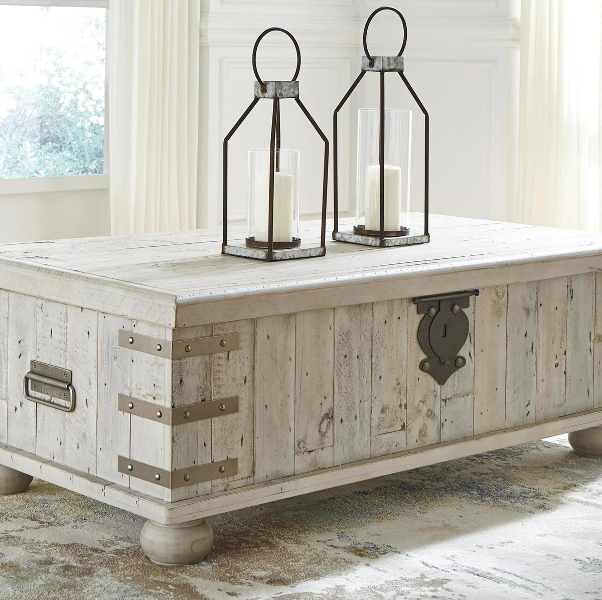 A rustic white-washed wooden coffee table styled as a storage chest, featuring distressed wood panels, metal corner brackets, and a decorative metal latch on the front. Two black metal lanterns with white candles are placed on top, set in a bright, neutral-toned room.