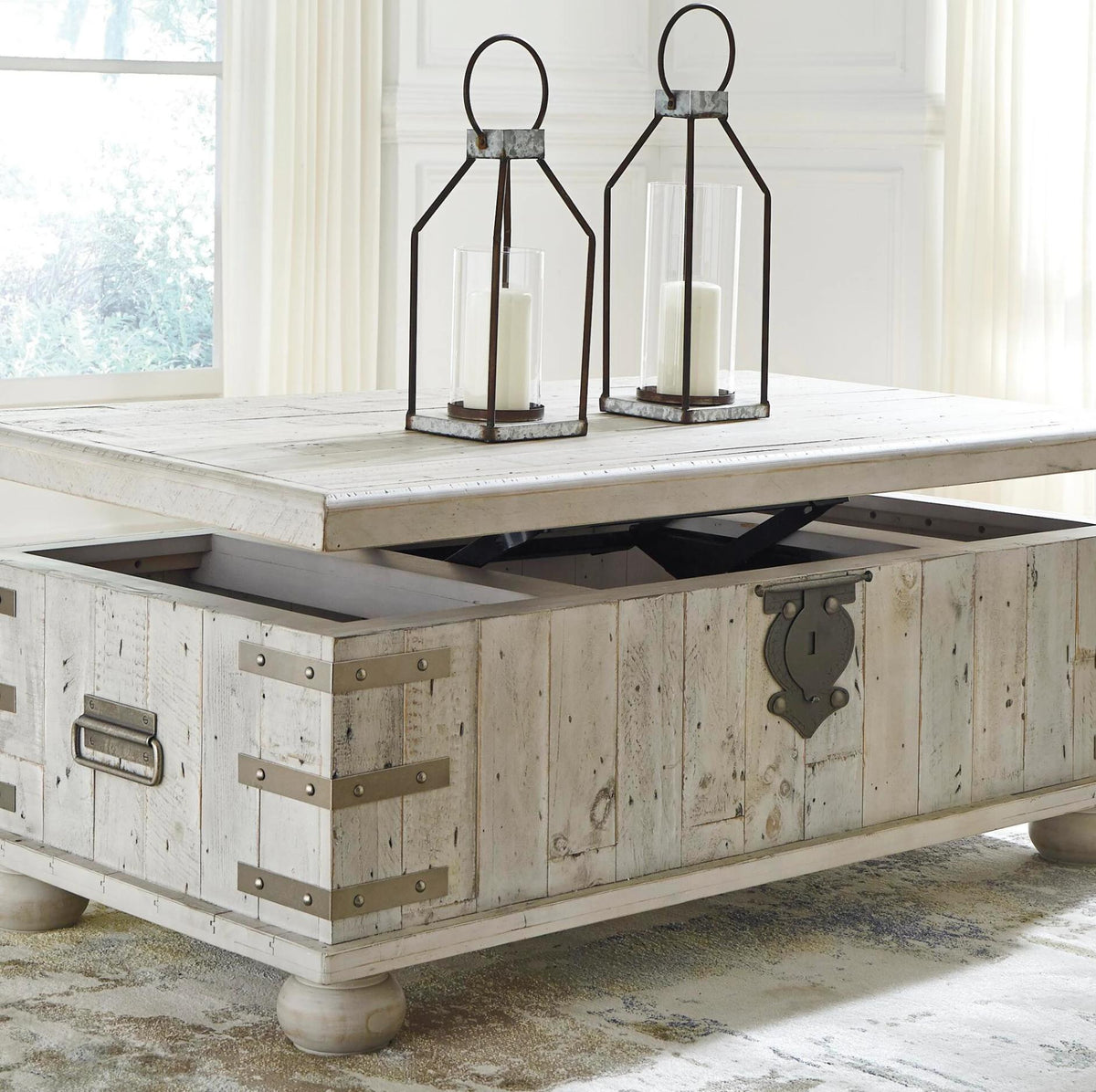 A rustic white-washed wood lift-top coffee table with an open storage compartment, styled with two black metal lantern candles on top in a bright room.