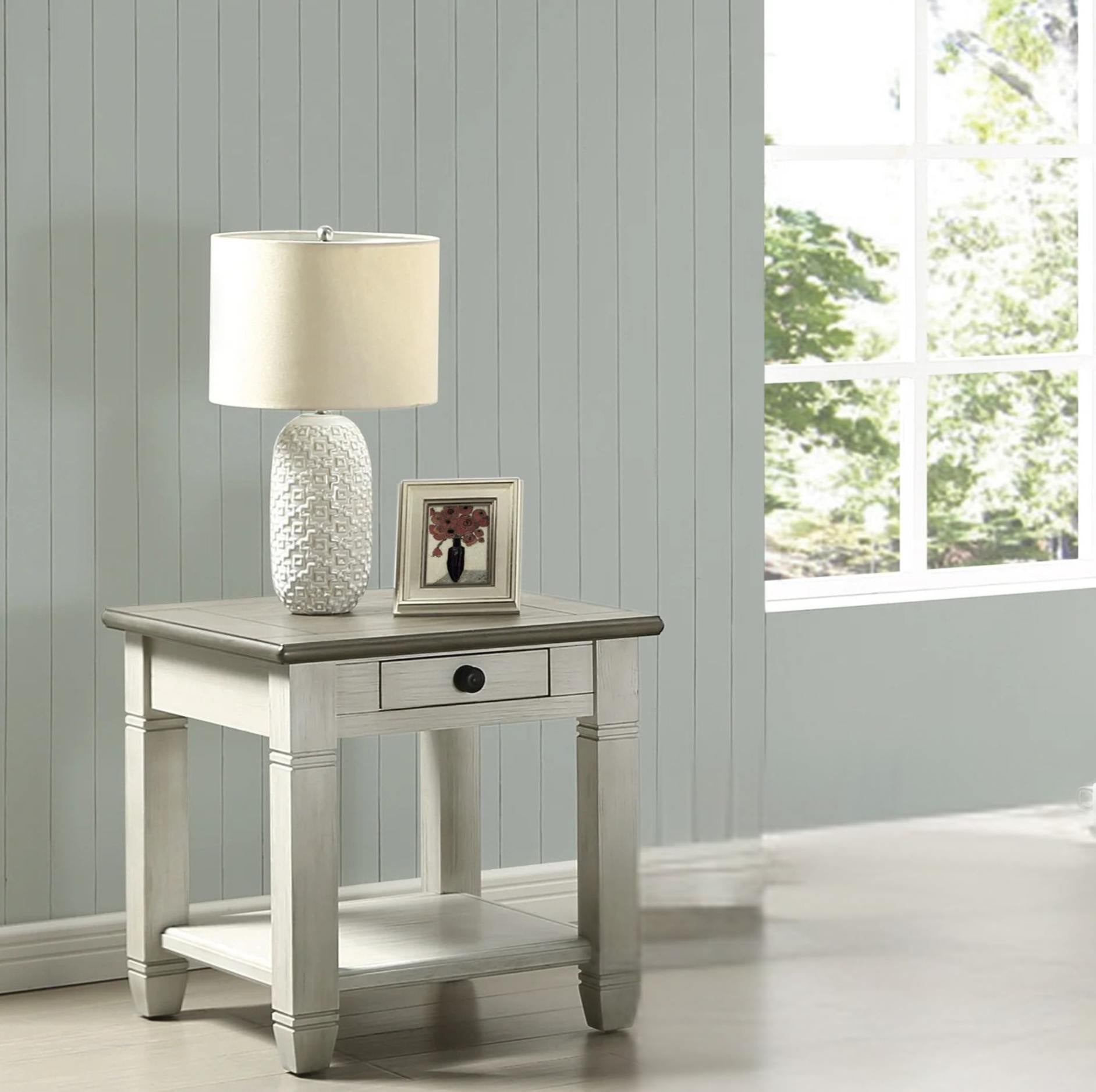 A light gray and white two-drawer end table styled with a textured white ceramic table lamp and a small picture frame, set against a light gray paneled wall next to a window.