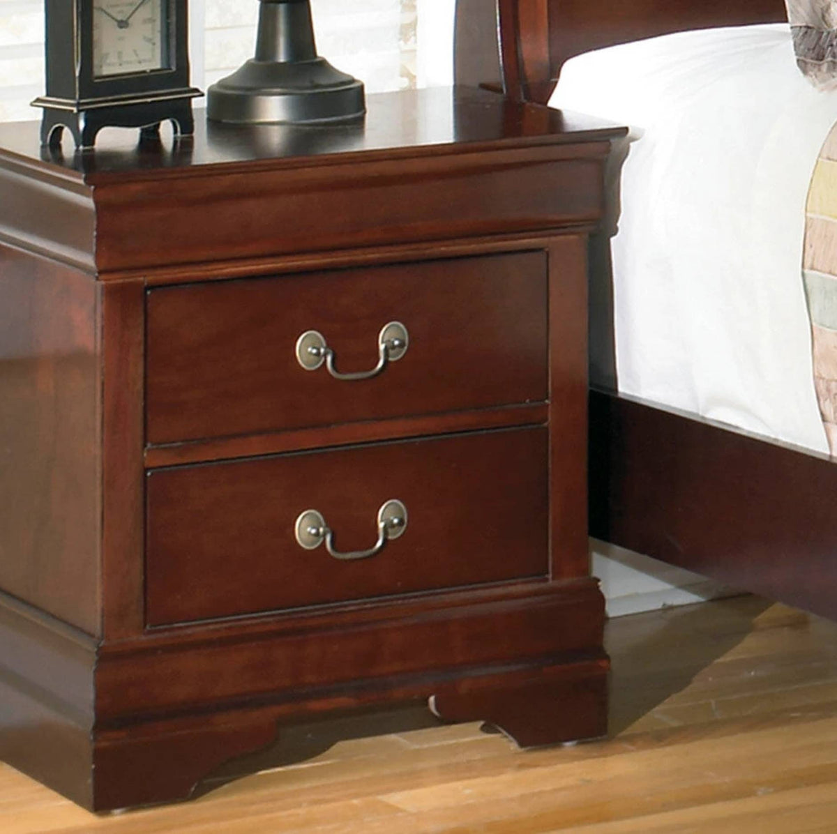 A dark reddish-brown wooden nightstand with two drawers and silver metal handles, shown next to a bed with white bedding and a lamp on top.