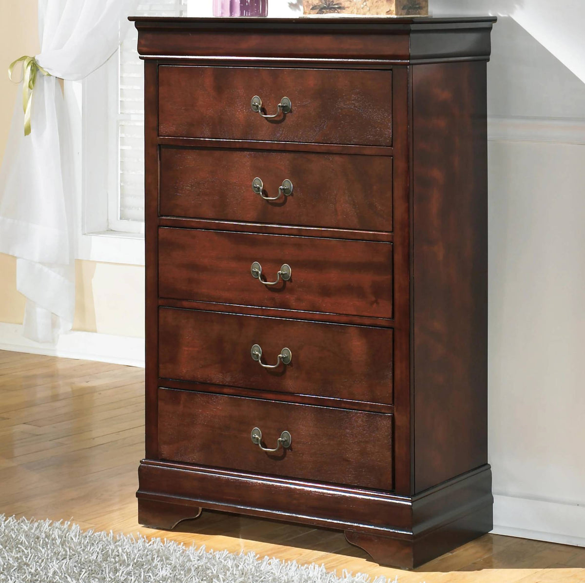 A five-drawer wooden chest of drawers in a warm reddish-brown finish, featuring dark metal pull handles, shown in a home interior setting.