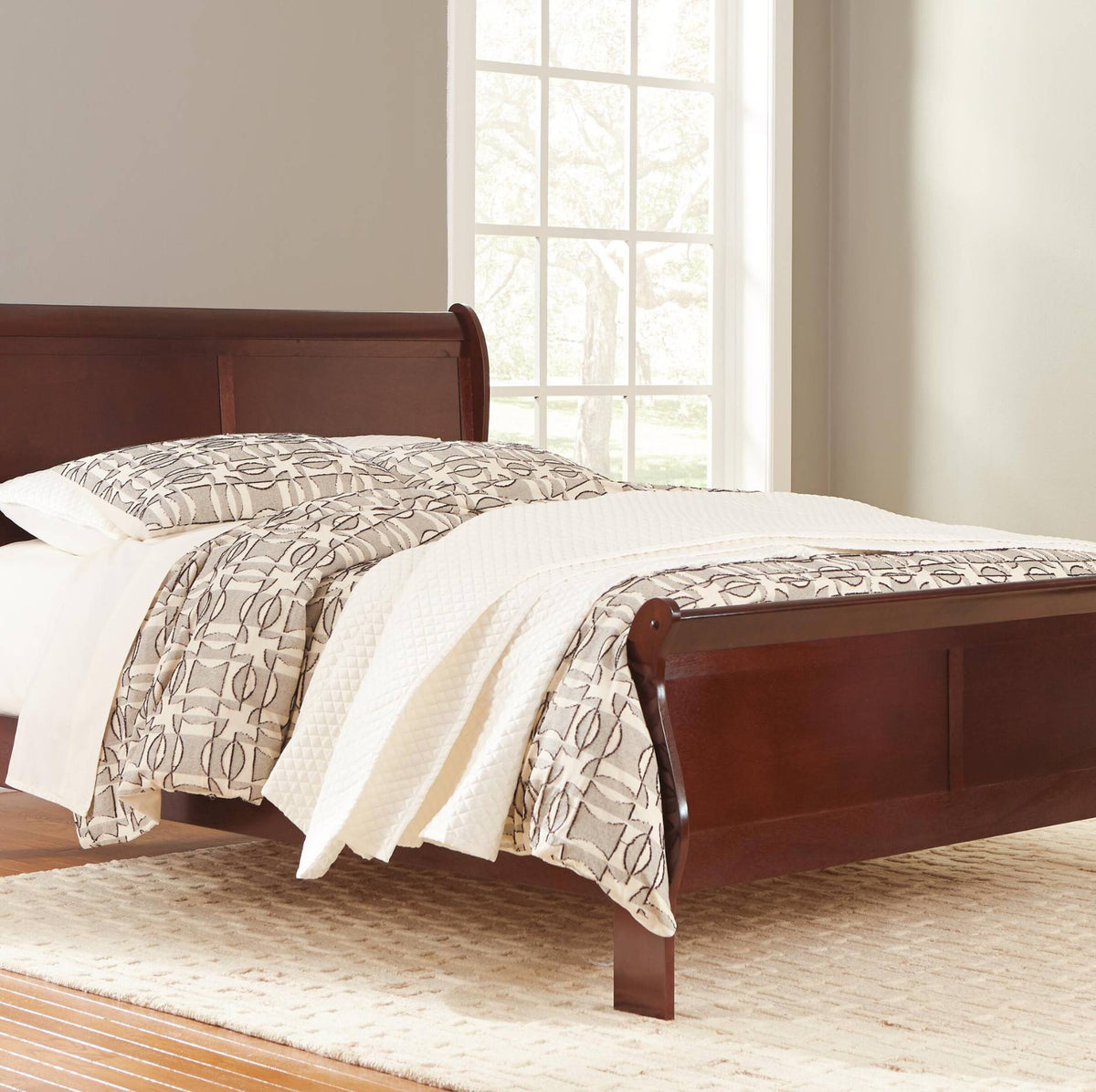 A dark reddish-brown wooden sleigh bed frame shown in a bedroom setting with patterned beige and white bedding, a window, and a light-colored rug.