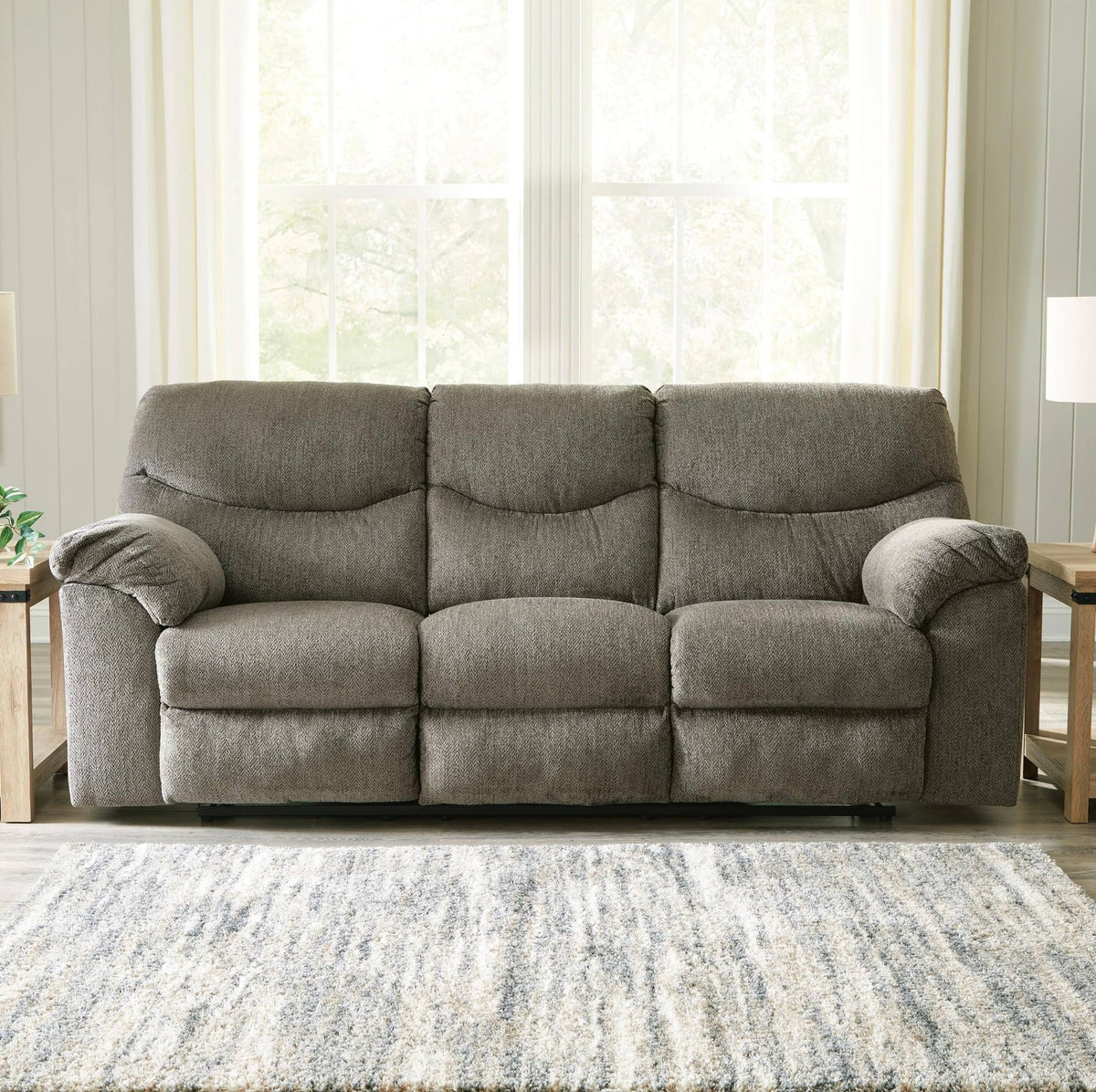 A three-seat gray reclining sofa with plush cushions, positioned in a bright living room setting with a patterned rug, side tables, and large windows.