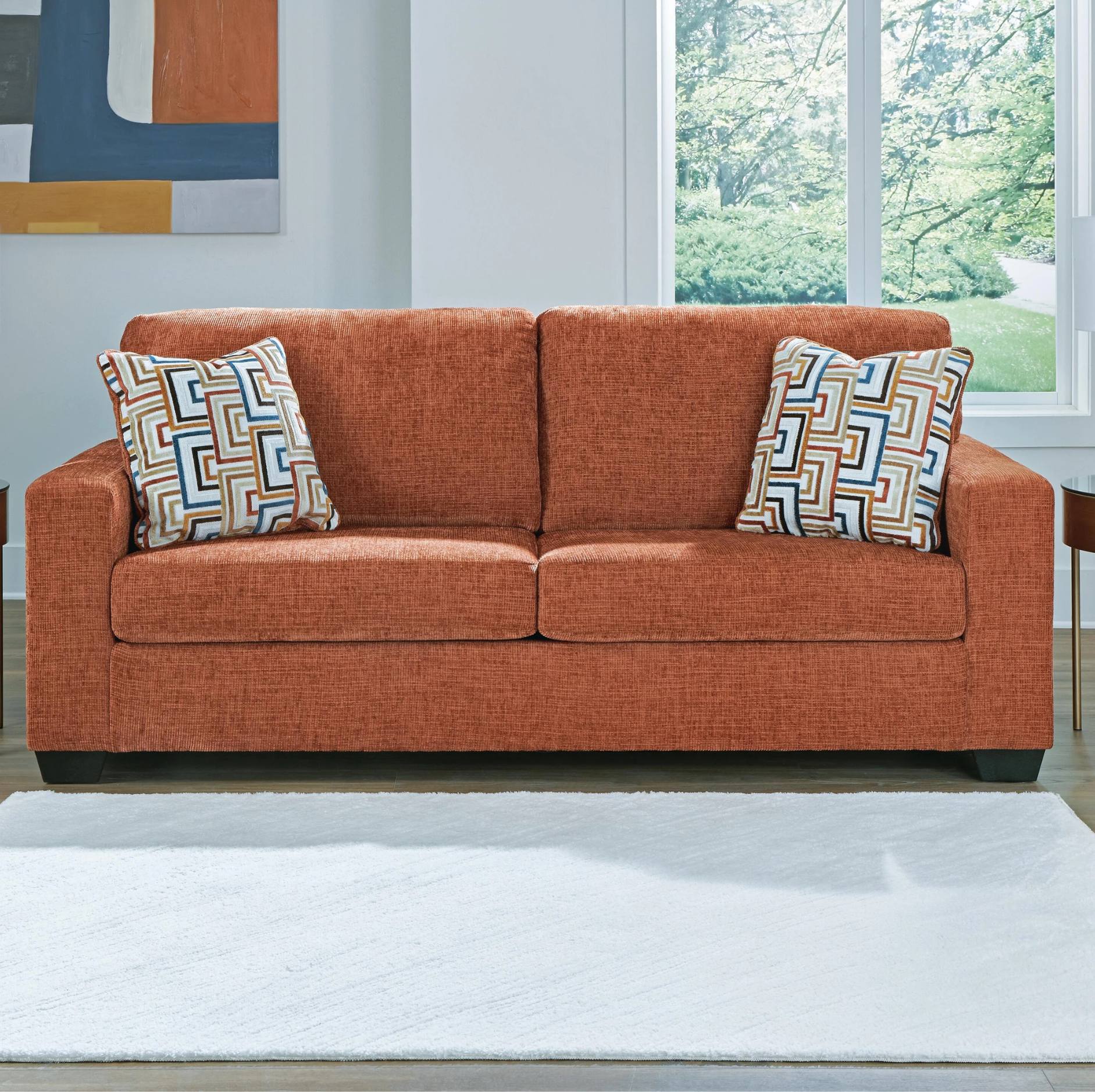 An orange-brown upholstered loveseat sofa with two patterned throw pillows, shown in a bright living room setting with a white rug, window, and wall art.