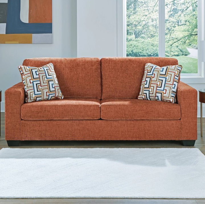 An orange-brown upholstered loveseat sofa with two patterned throw pillows, shown in a bright living room setting with a white rug, window, and wall art.