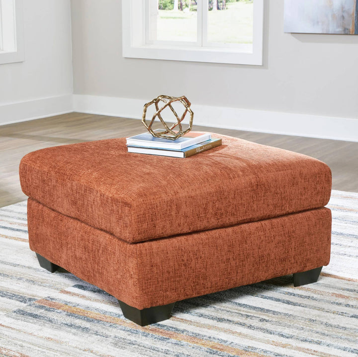 A square upholstered ottoman in a textured rust-orange fabric, styled with two books and a decorative gold geometric sculpture on top, set in a bright room on a striped rug.