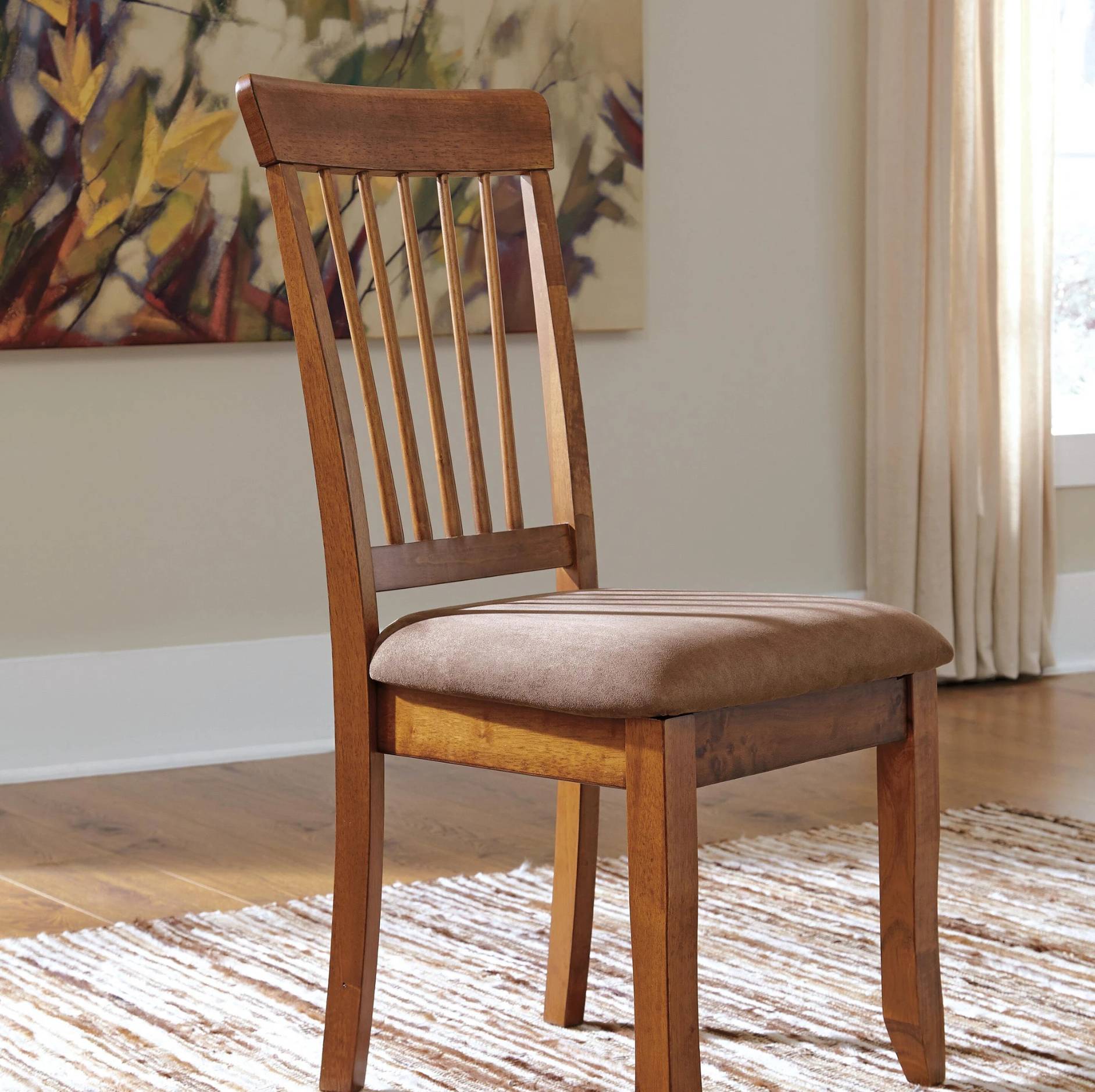 A wooden dining chair with a slatted backrest and a brown upholstered seat cushion, shown in a real interior room with a rug, curtains, wall art, and wood floor.