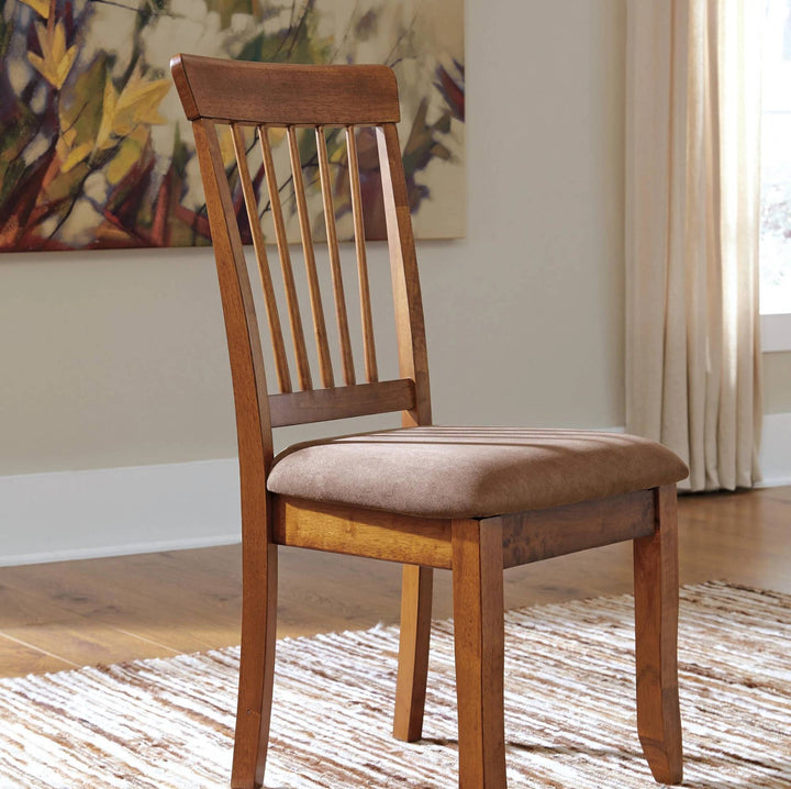 A wooden dining chair with a slatted backrest and a brown upholstered seat cushion, shown in a real interior room with a rug, curtains, wall art, and wood floor.