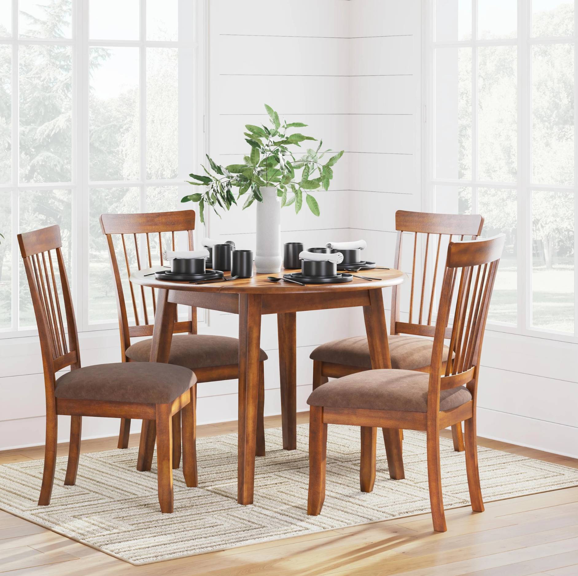 A 5-piece wooden dining table set with four slat-back chairs featuring brown upholstered seats, shown in a bright dining room setting with a vase of green leaves and black cups on the table.
