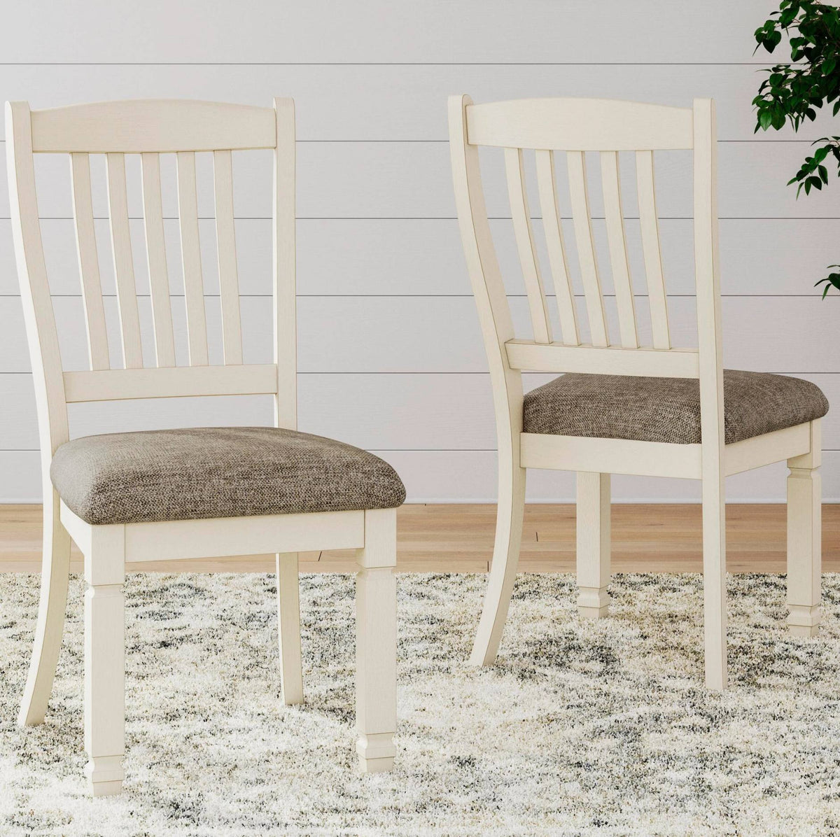 Two cream dining chairs with upholstered gray-brown seat cushions, shown on a shag rug against a white paneled wall.