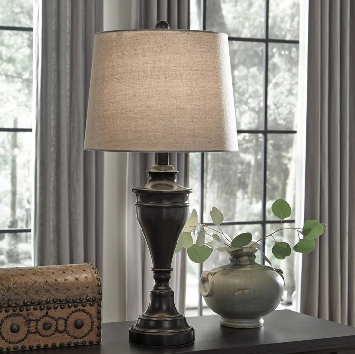 A table lamp with a dark bronze-finished pedestal base and a beige linen drum shade, shown on a dark tabletop in a styled interior setting with a window, curtains, a decorative box, and a small vase with greenery.