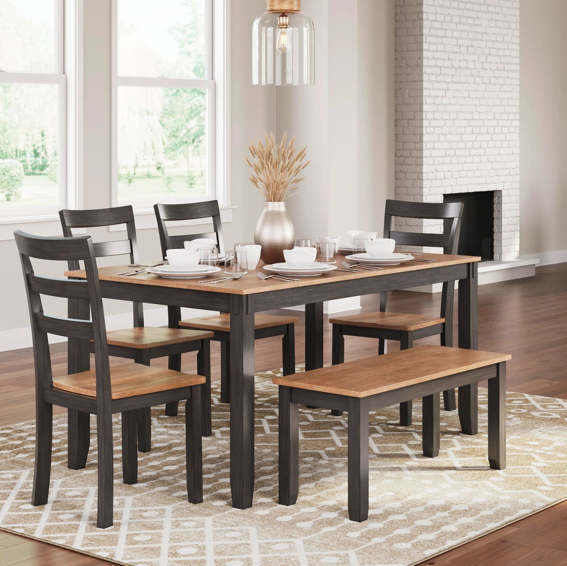 A 6-piece dining room set featuring a rectangular table with a medium brown wood top and dark charcoal gray legs, accompanied by four matching ladder-back chairs, a long bench, and a decorative vase on a patterned area rug in a bright dining room.