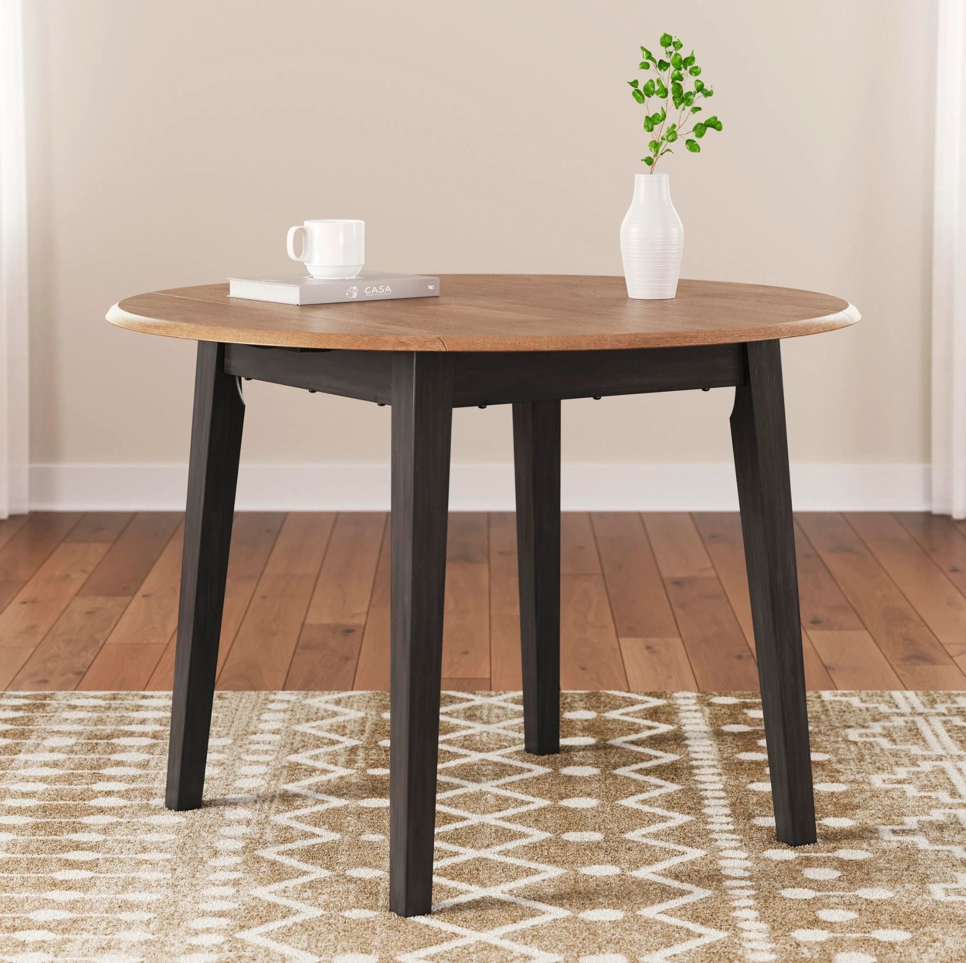 A round wooden dining table with dark black legs, styled with a white mug, a book, and a small white vase with green leaves on a patterned rug in a room with wood floors and beige walls.
