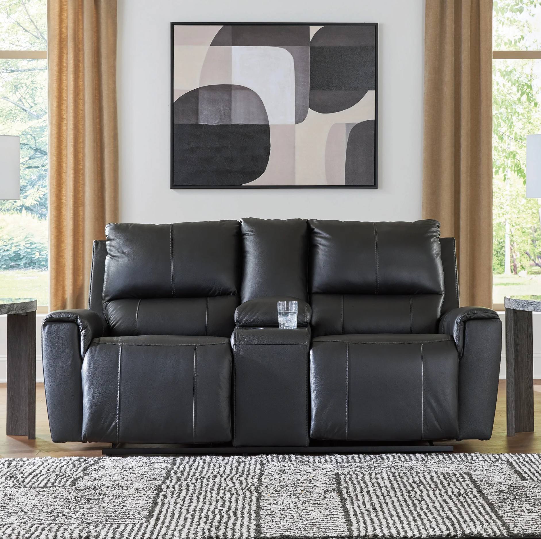 A black leather reclining loveseat sofa centered in a staged living room setting, with a matching armchair partially visible on both sides, a patterned rug in front, and abstract wall art above.