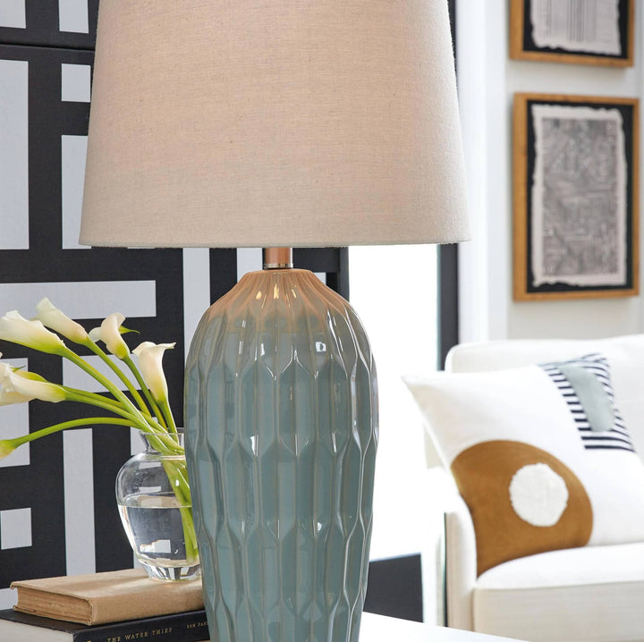 A table lamp with a textured, faceted ceramic base in a muted blue-gray finish and a beige fabric drum shade, styled on a table next to a vase of white calla lilies.