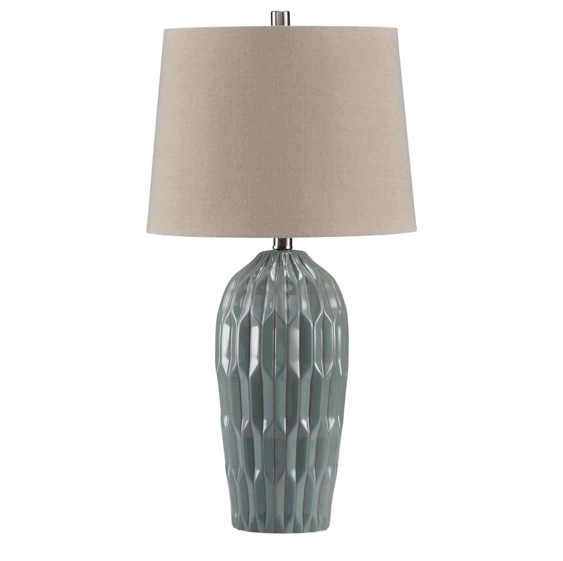 A table lamp with a textured, faceted ceramic base in a muted sage green-gray color and a tapered drum shade in a warm beige fabric, shown against a plain white background.