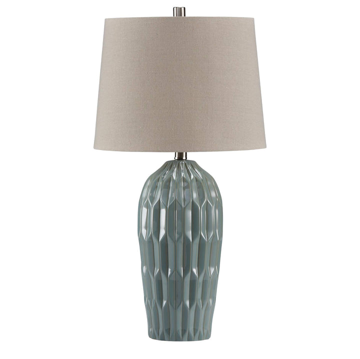 A table lamp with a textured, faceted ceramic base in a muted sage green-gray color and a tapered drum shade in a warm beige fabric, shown against a plain white background.