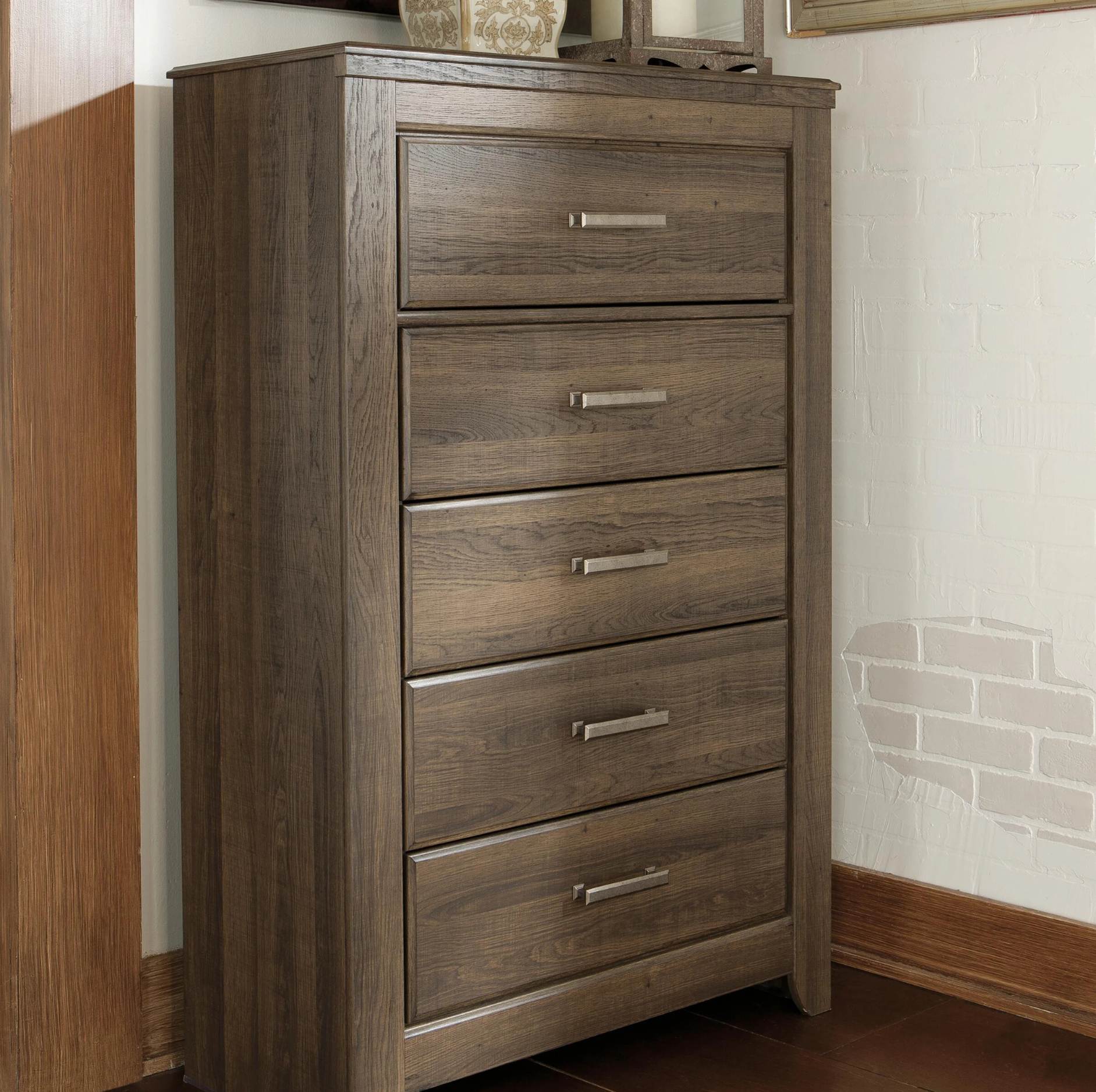 A five-drawer wooden chest of drawers with a weathered brown finish and silver metal handles, shown in a room corner next to a wood-paneled wall.