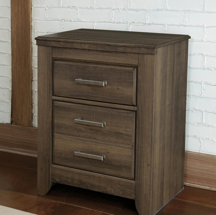 A two-drawer nightstand in a medium brown wood finish with silver metal handles, set against a white brick wall and wooden floor.