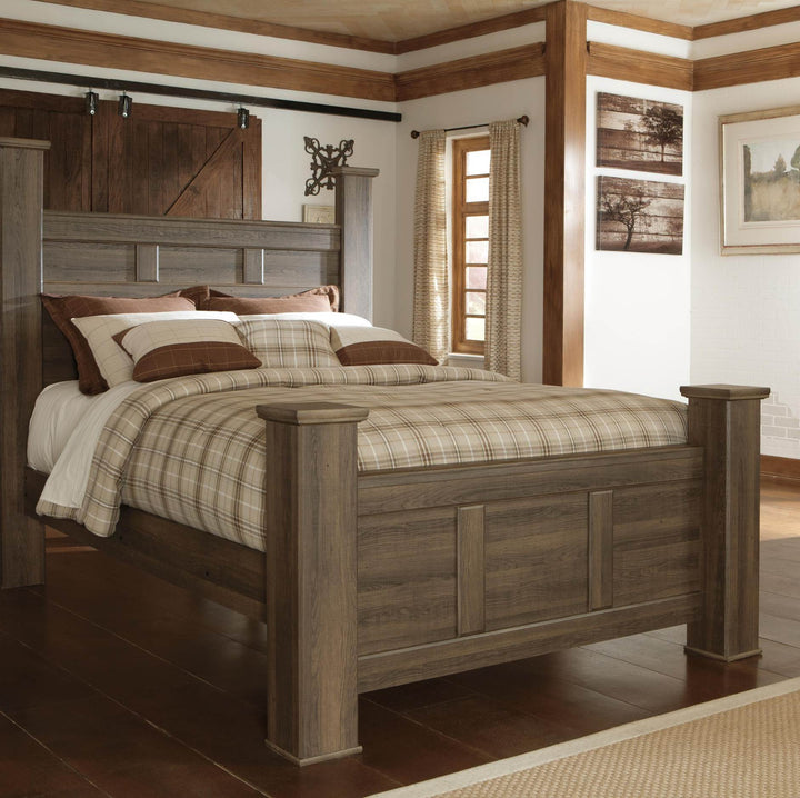 A rustic wooden platform bed with a weathered gray-brown finish, styled with a beige and brown plaid comforter and white pillows, set in a cabin-style bedroom with wood-paneled walls and a window.