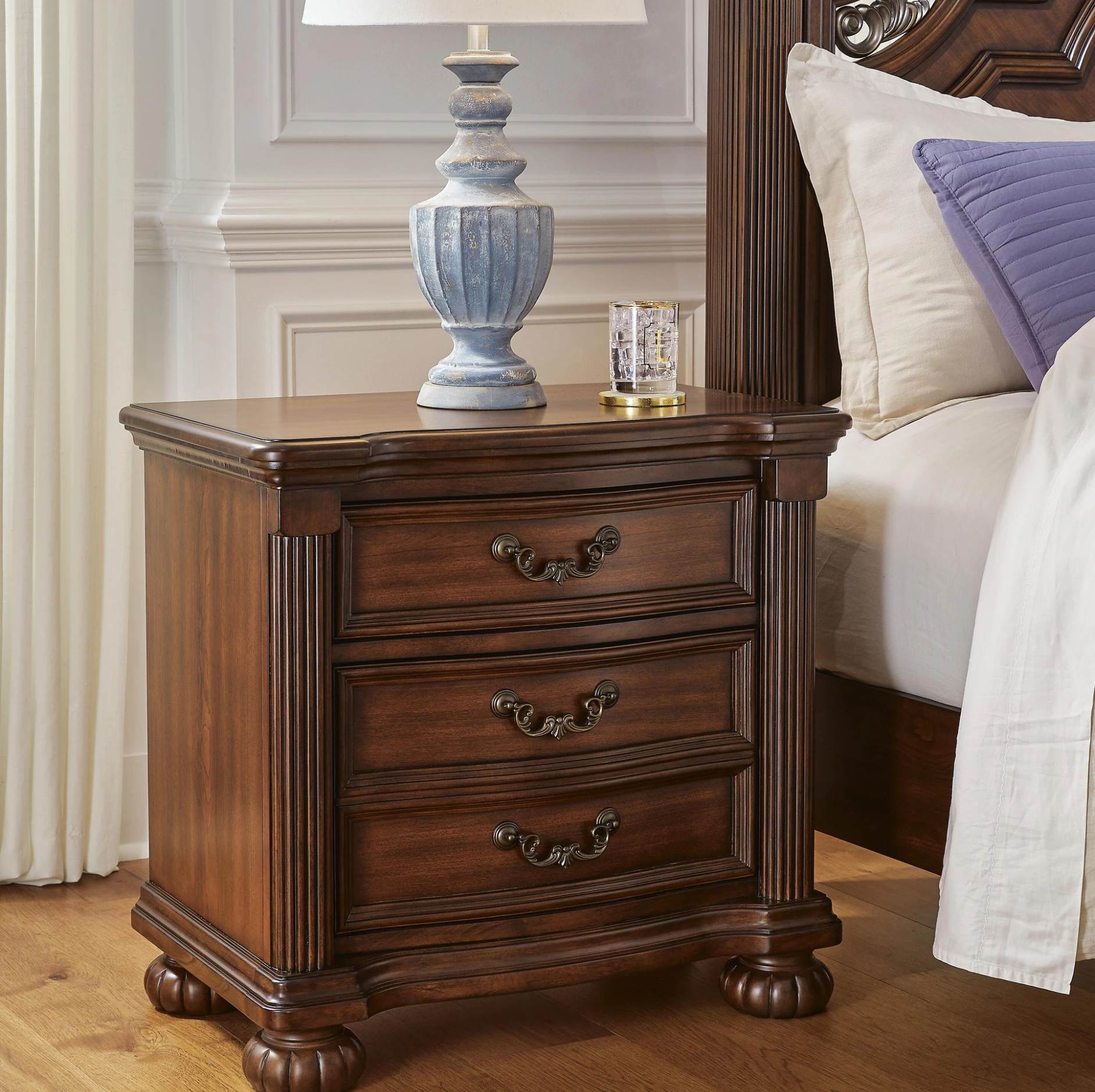 A dark brown wooden nightstand with three drawers and ornate metal handles, styled next to a bed with white and purple pillows. A blue ceramic table lamp and a small glass candle holder sit on top of the nightstand.