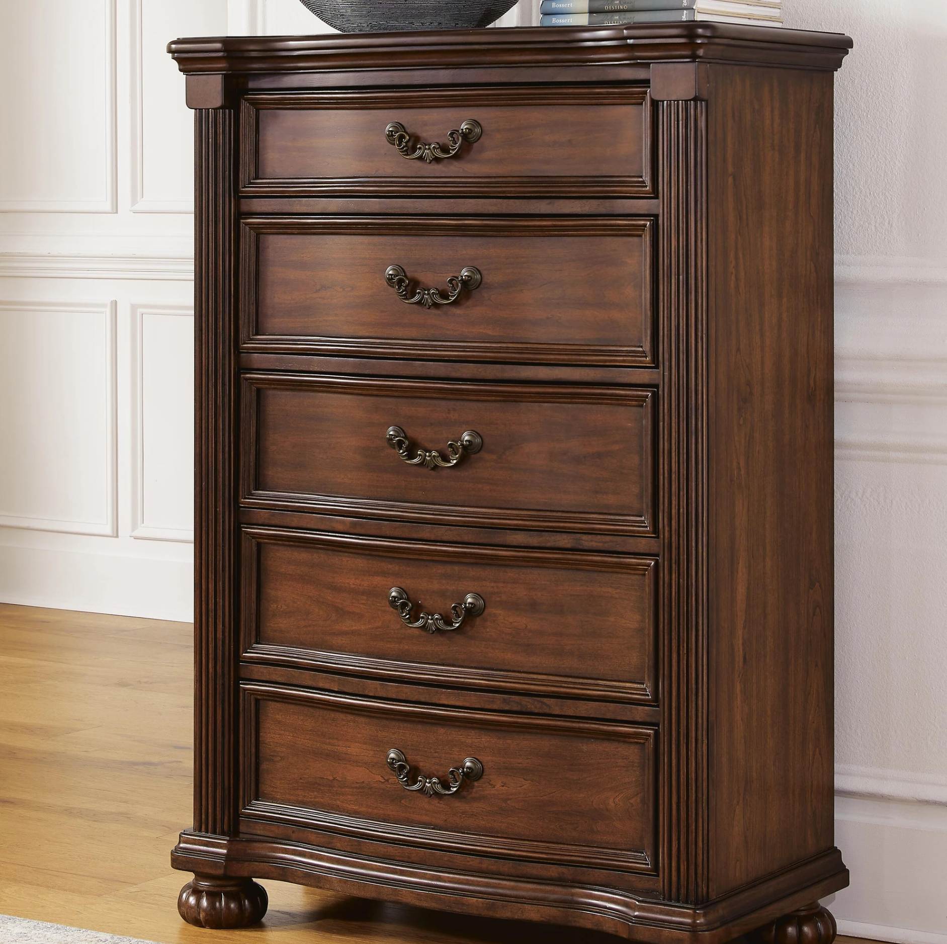 A dark brown wooden five-drawer chest of drawers with ornate metal handles, set against a white paneled wall and light wood floor.