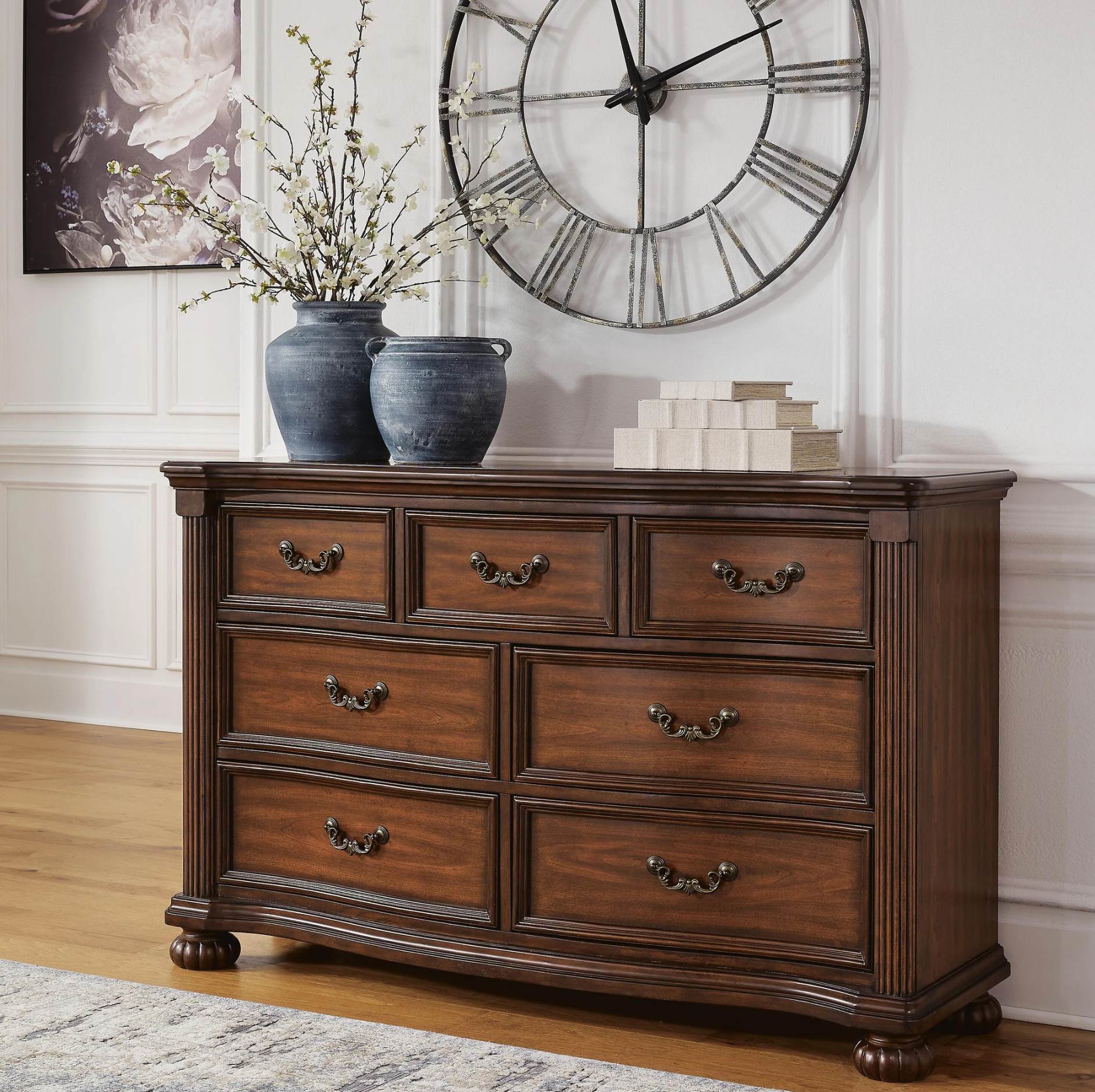 A dark brown wooden dresser with eight drawers and ornate metal pulls, styled in a living room setting with blue vases, a large clock, and a stack of books on top.