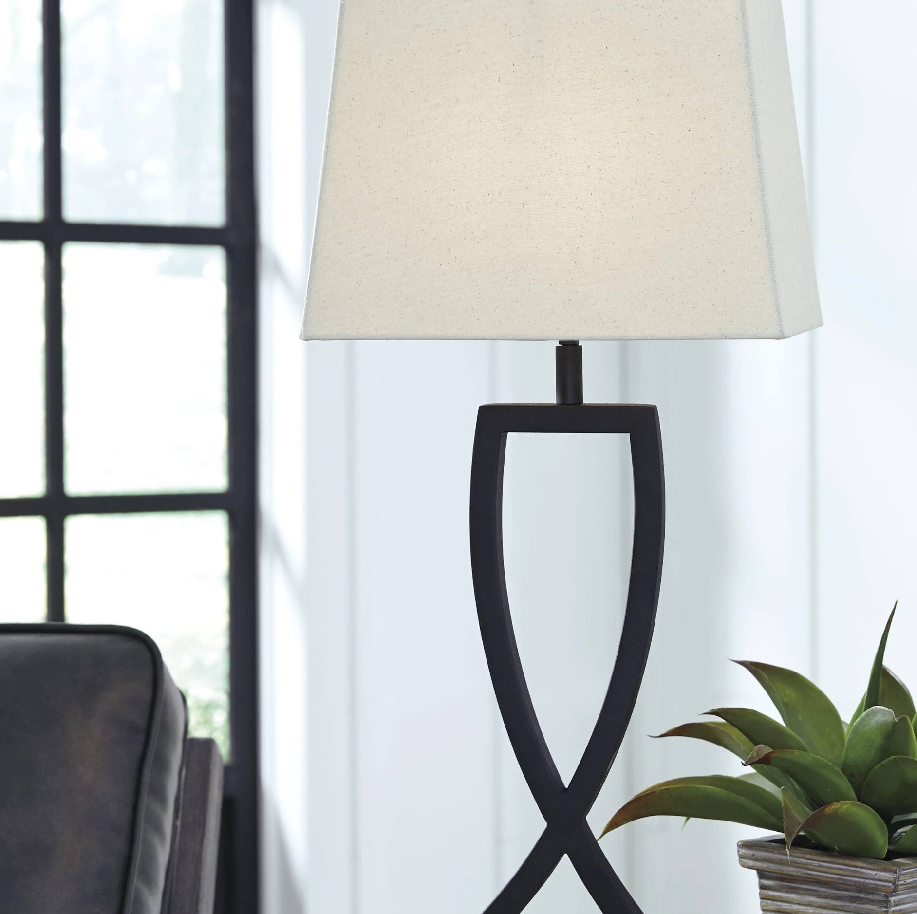 A table lamp with a black sculptural base and a cream fabric drum shade, shown lit in a bright interior setting with a window, chair, and potted plant.