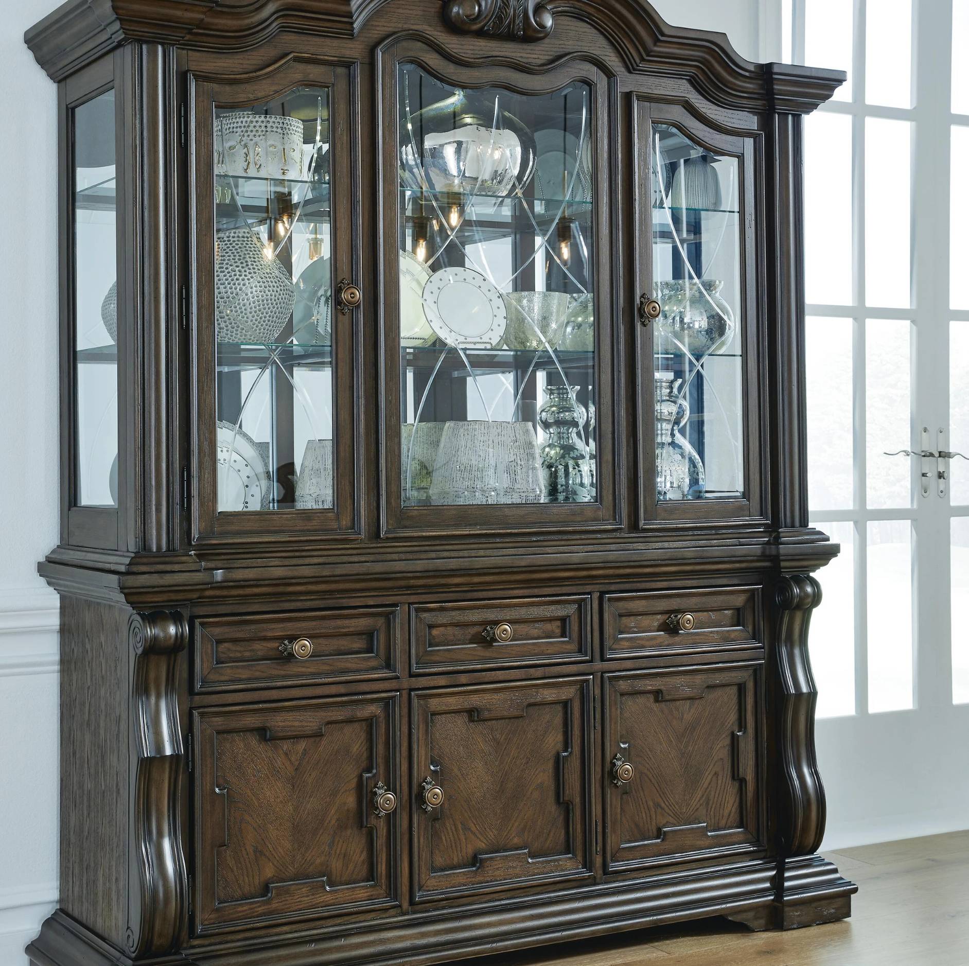 A large dark wood hutch or china cabinet with glass-front upper doors, curved top molding, lower drawers, and paneled cabinet doors, shown in a bright room with white walls and a window.
