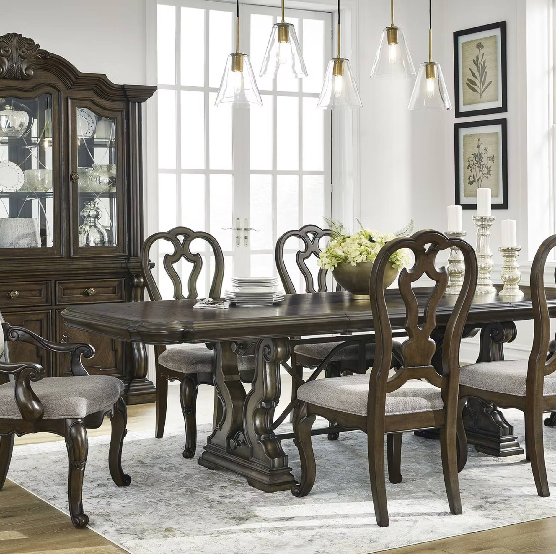 A dark wood dining room set including a rectangular table, six matching chairs with upholstered seats, and a matching hutch, arranged in a bright dining room with pendant lights, a patterned rug, and framed botanical prints.
