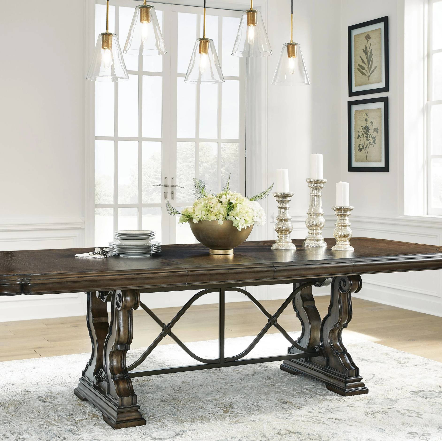 A dark wood dining table with ornate carved legs and a central metal stretcher bar, styled in a bright dining room with pendant lights, candles, and a floral centerpiece.