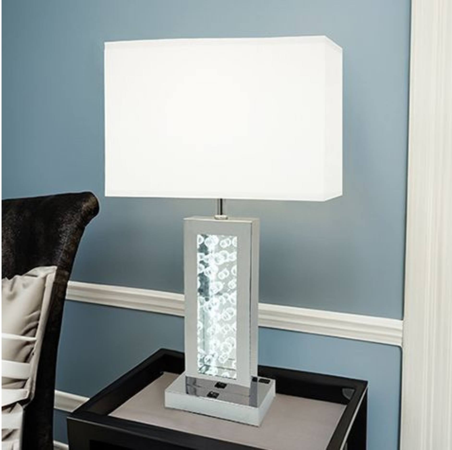 A modern table lamp with a white rectangular fabric shade, a clear faceted crystal column, and a silver-toned base, shown on a dark side table against a blue wall.