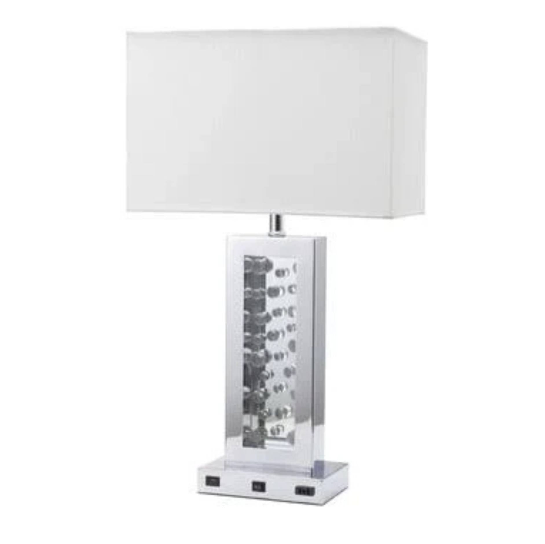 A modern table lamp with a white rectangular fabric shade, a clear glass column containing stacked silver and black LED light strips, and a square chrome base with built-in power outlets.