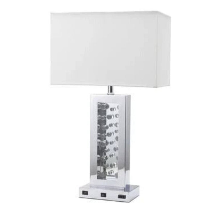 A modern table lamp with a white rectangular fabric shade, a clear glass column containing stacked silver and black LED light strips, and a square chrome base with built-in power outlets.