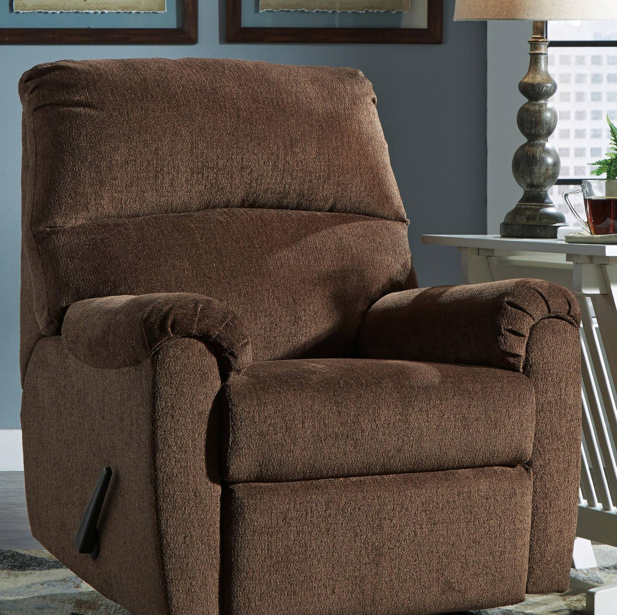 A brown textured fabric recliner chair shown in a living room setting with a side table, lamp, and wall visible.