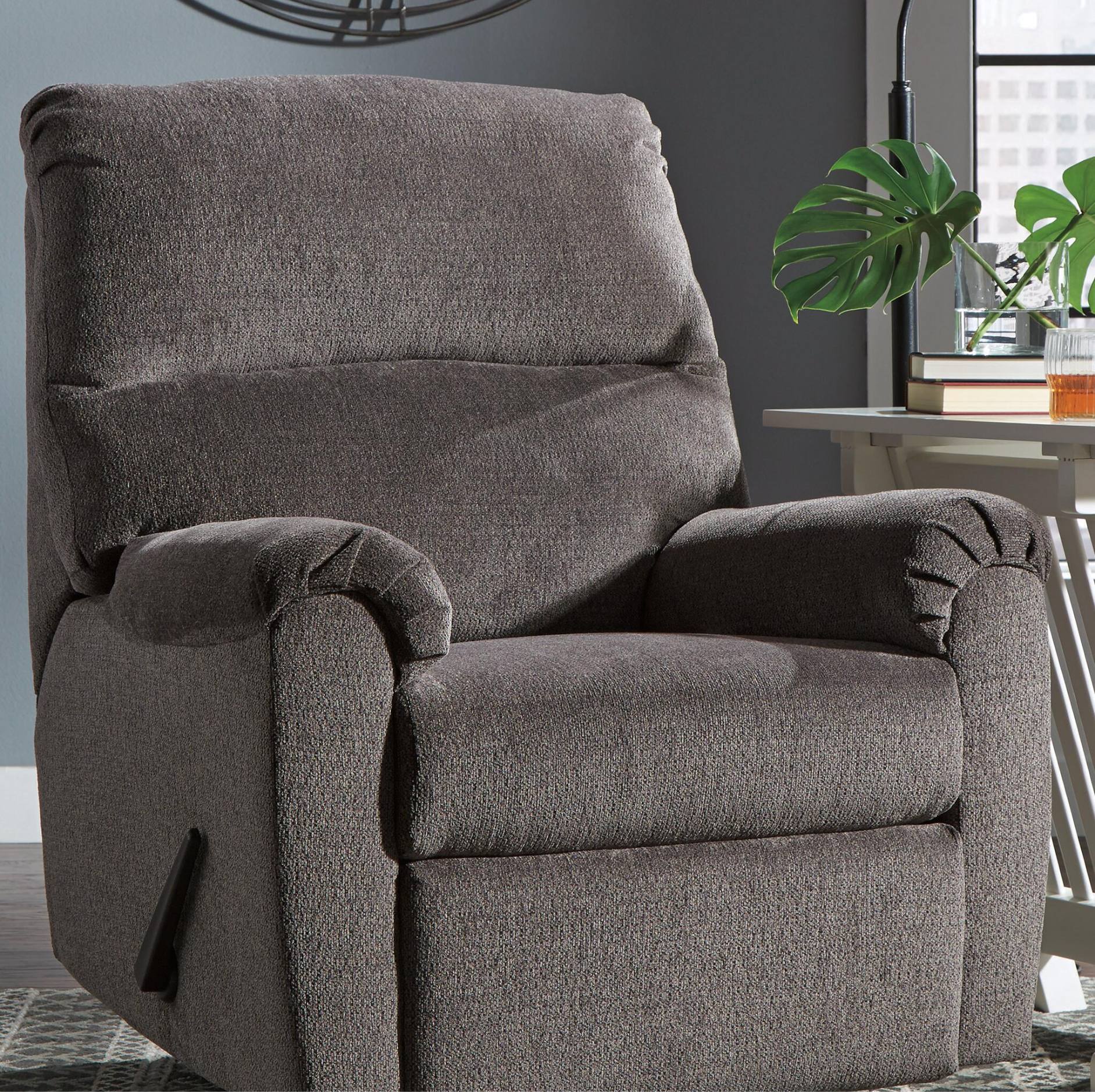 A plush upholstered recliner chair in dark taupe-brown textured fabric, shown in a modern living room setting with a side table, plant, and window in the background.