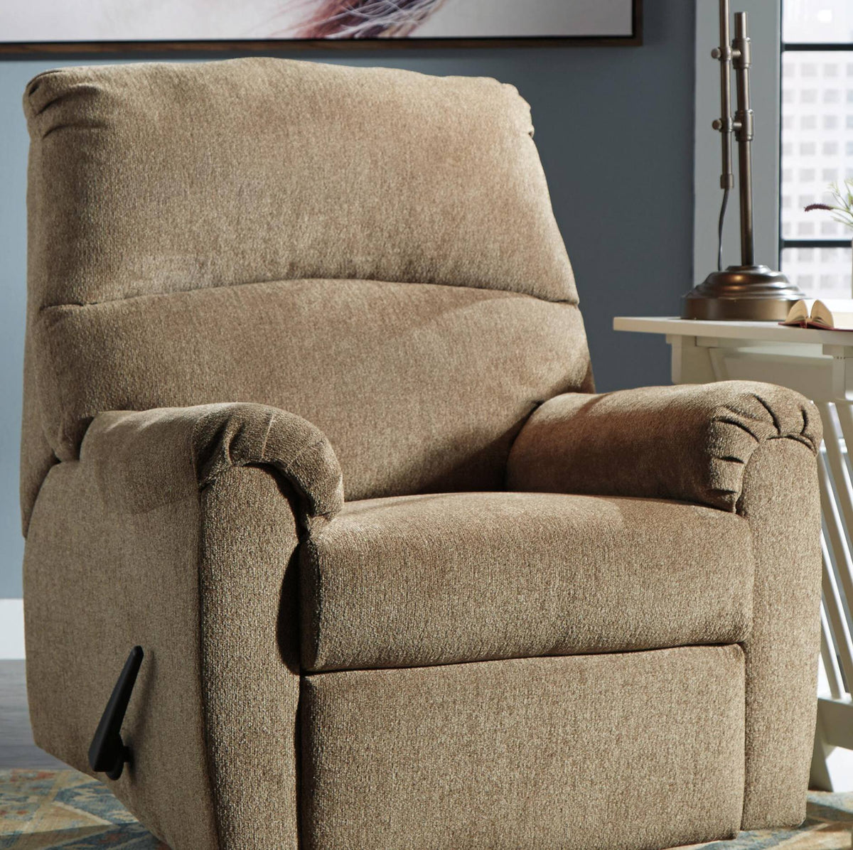 A plush upholstered recliner chair in a warm taupe-beige fabric, shown in a real living room setting with a side table, lamp, window, and wall art visible.