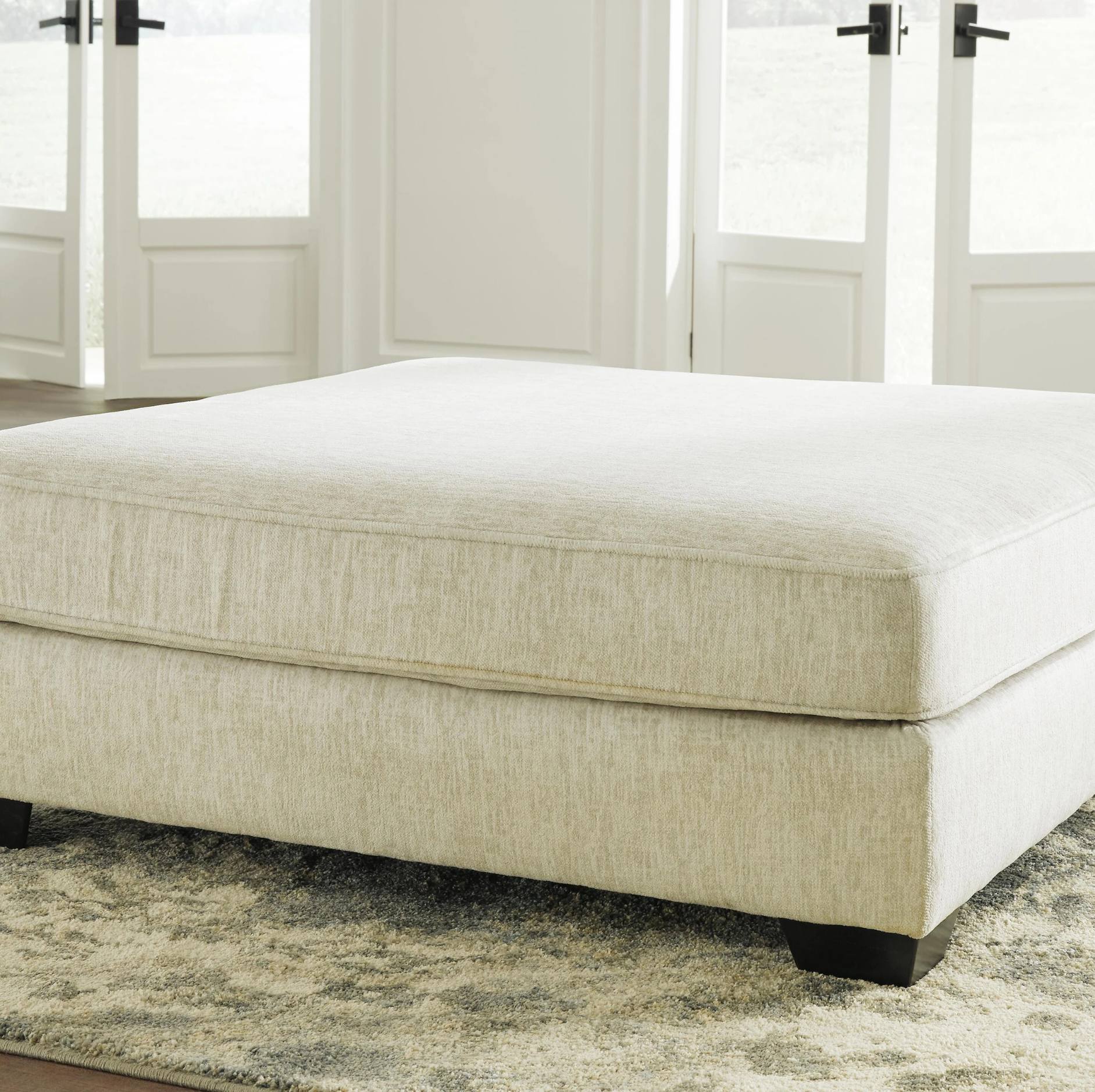 A large upholstered ottoman or low bench in a light beige textured fabric, shown in a bright room with white French doors and a patterned rug.