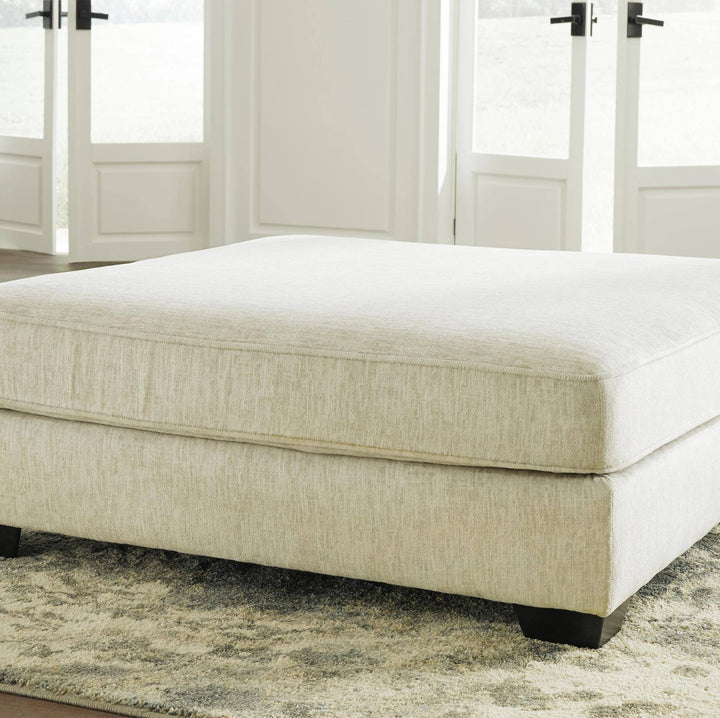 A large upholstered ottoman or low bench in a light beige textured fabric, shown in a bright room with white French doors and a patterned rug.