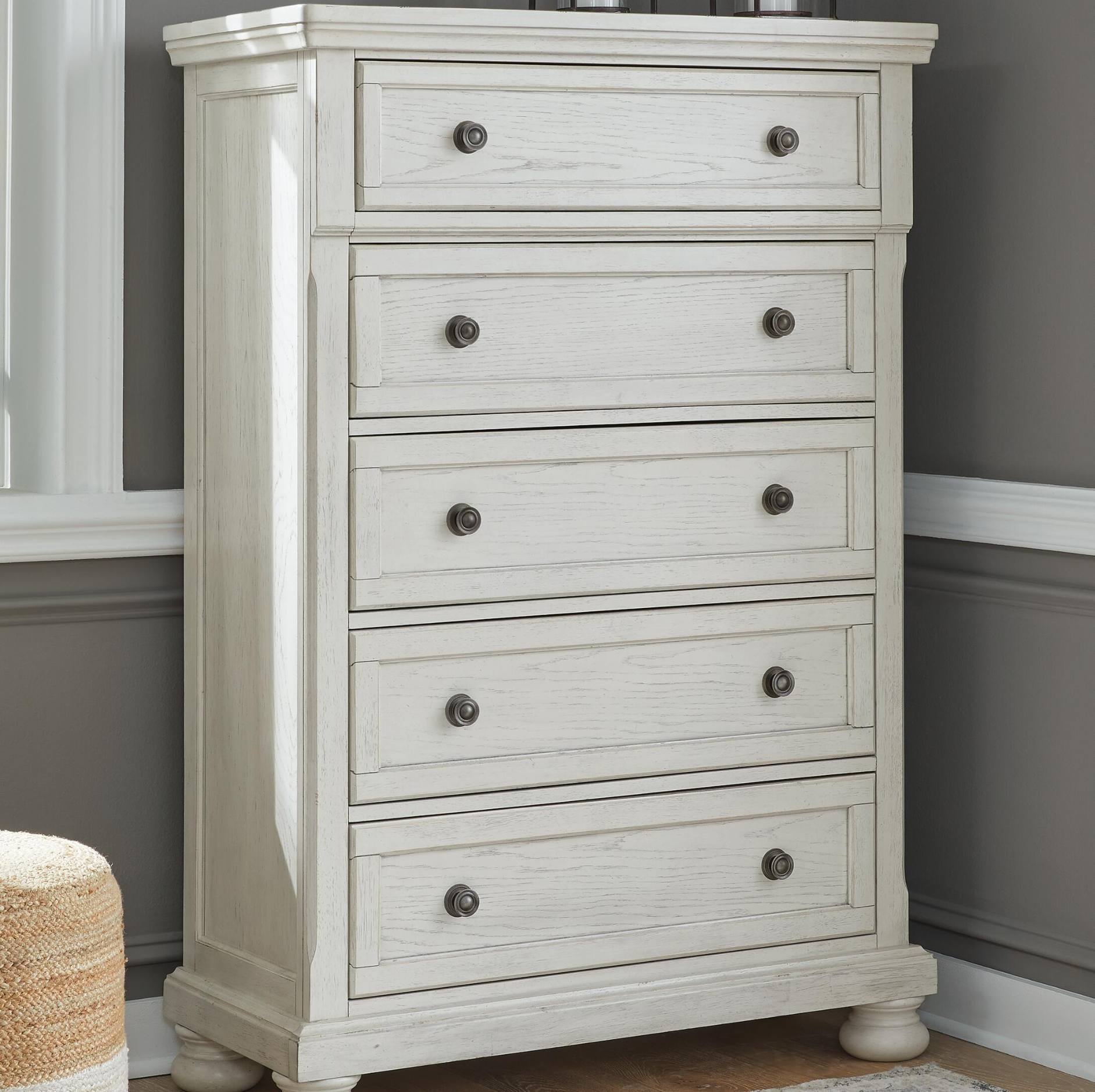 A five-drawer chest of drawers in a distressed white or off-white finish, featuring round dark metal knobs and turned feet, set against a gray wall with white trim.