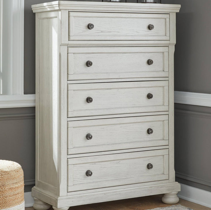 A five-drawer chest of drawers in a distressed white or off-white finish, featuring round dark metal knobs and turned feet, set against a gray wall with white trim.