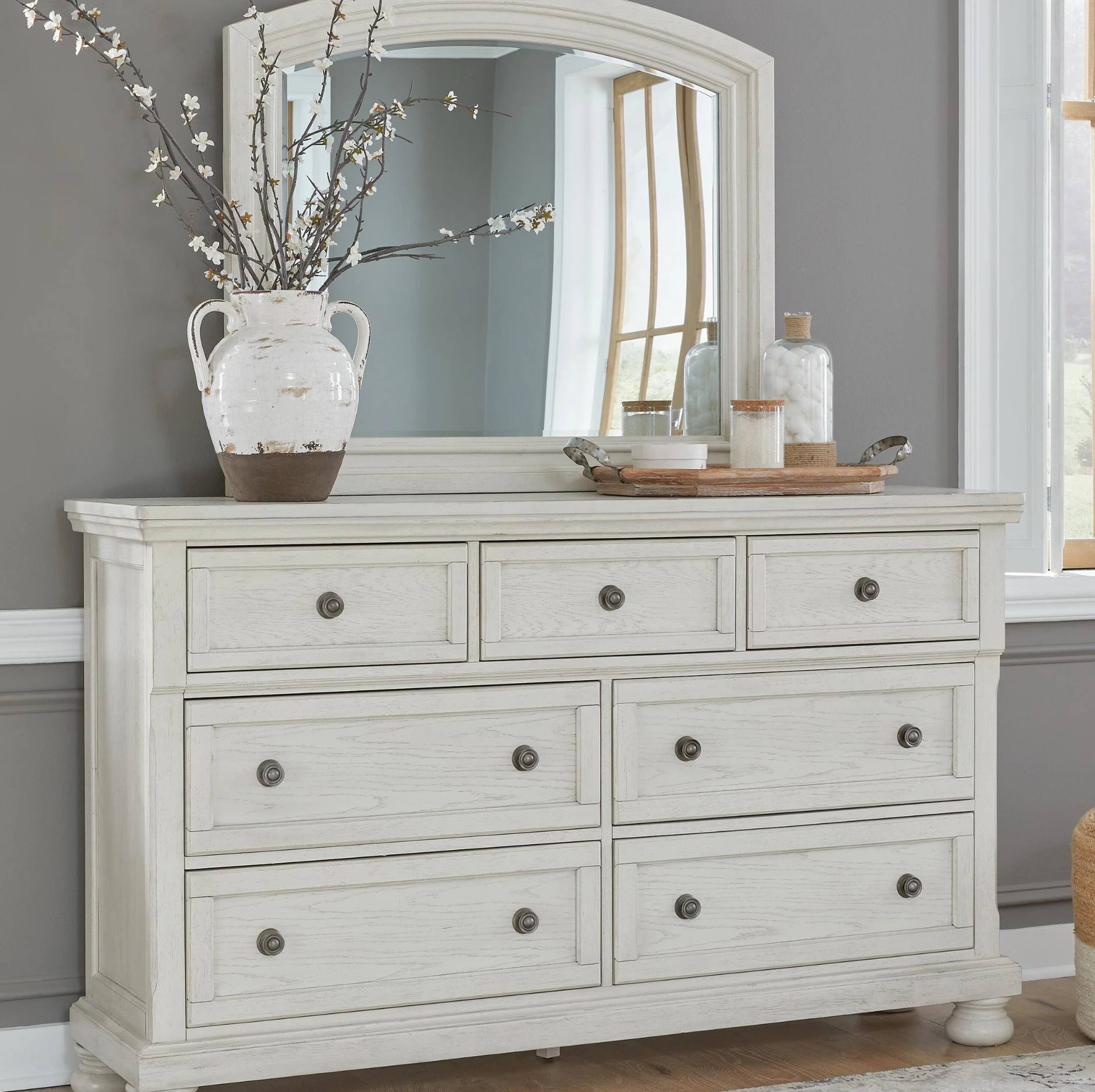 A distressed white eight-drawer dresser with dark round knobs, topped with a matching arched mirror. The dresser is styled in a bedroom setting with a large ceramic vase of branches on top, alongside decorative bottles and a tray.