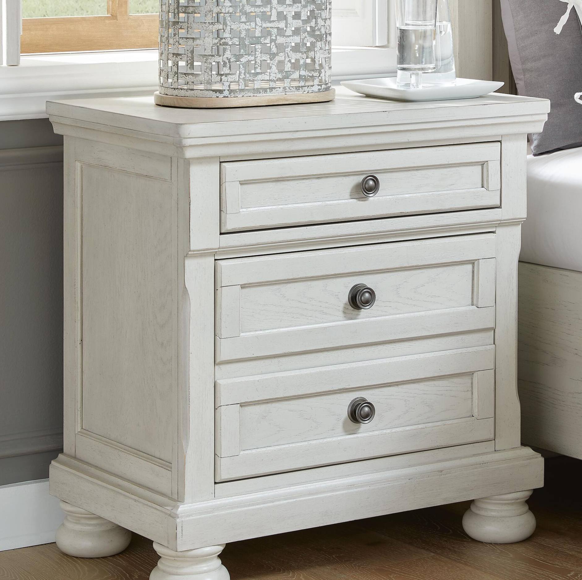 An off-white three-drawer nightstand with silver metal knobs, shown in a bedroom setting next to a window and bed.