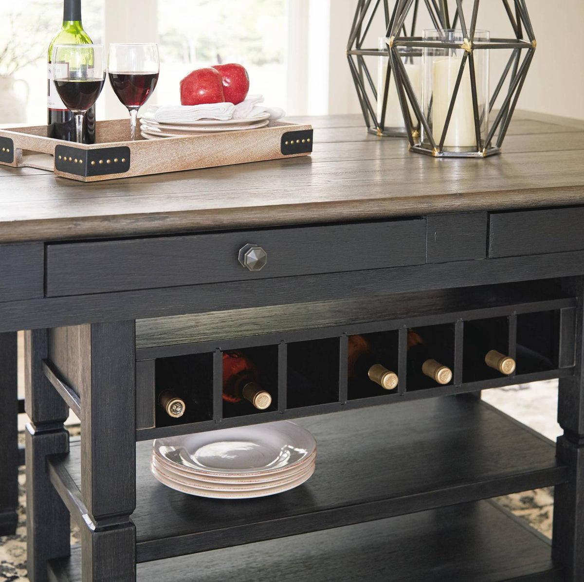 A dark charcoal gray wood wine bar console table with a rustic light brown wood top, featuring a single drawer, built-in wine bottle storage, and a lower shelf holding plates. The table is styled with a wine tray, glasses of red wine, a bottle, and a geometric metal candle holder against a bright, softly blurred background.