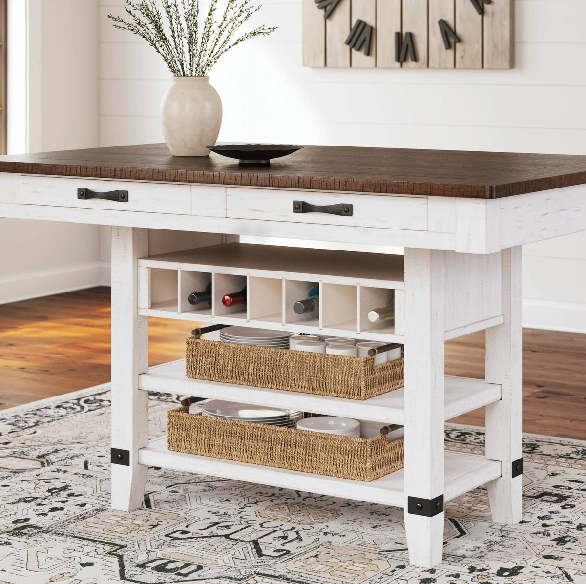 A white kitchen island table with a dark wood top, three drawers with black handles, open shelving, two woven wicker baskets, and black metal bracket accents on the legs. The table is styled in a bright kitchen with a vase of branches and a plate on top.